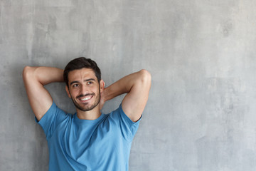 Handsome smiling young man in blue t-shirt leaning against gray textured wall with hands behind head, copy space for your text