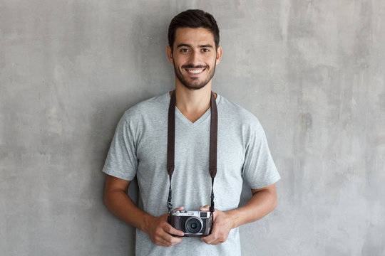 Smiling handsome male photographer in t shirt, holding retro film photo camera, standing against gray textured wall