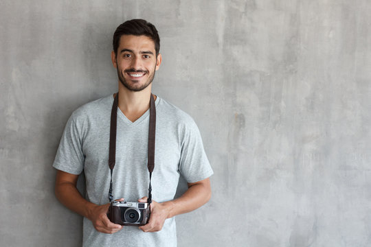 Portrait of smiling handsome male photographer in gray t shirt, holding his camera, standing against gray textured wall with copy space for your ads