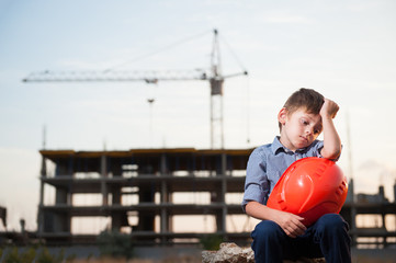 cute upset sad little boy sitting near building site with orange helmet in hands