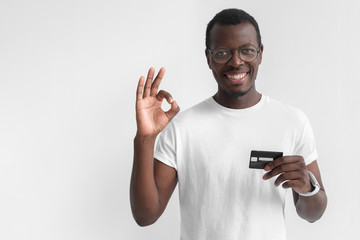 Young smiling dark skin african man in white t shirt holding credit card and showing okay sign...