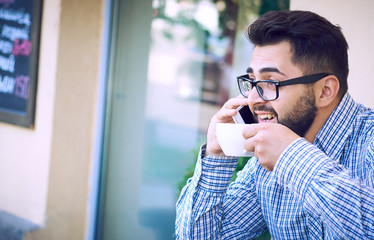 Modern hipster businessman with beard and glasses drinking coffee in the city cafe during lunch time and using mobile phone.