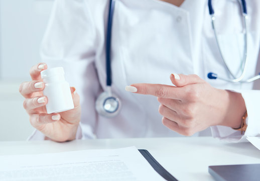 Young female medicine doctor sitting in front of working table holding jar of pills and explains to the patient how to take medication. Medical and pharmacy concept