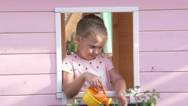 Little girl watering flowers in window box on pink playhouse.