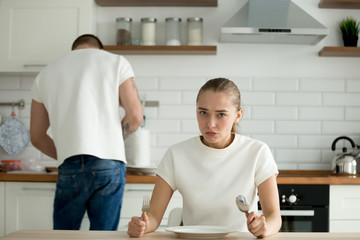 Hungry wife sit at table dissatisfied with man cooking dinner, funny impatient mad woman waiting food serving prepared by boyfriend, husband slave over hot stove. Concept of gender equality, feminism