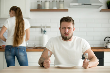 Dissatisfied hungry male impatiently waiting for dinner, mad man sitting at table with empty plate want to eat, busy wife prepare food for husband cooking dishes in kitchen. Gender inequality concept