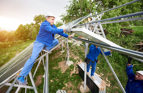 Installing of stand-alone solar photo voltaic panel system. Two technicians in hard-hats mounting big shiny solar module on platform on green summer view background. Alternative energy concept.