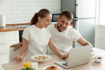 Happy millennial couple enjoy healthy homemade breakfast at home together reading funny news at laptop, smiling man and woman eat tasty delicious porridge or oatmeal browsing internet on computer