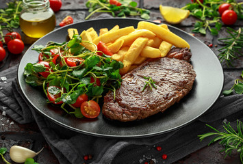 Grilled sirloin steak with potato fries and vegetables, tomato salad in a black plate. rustic table