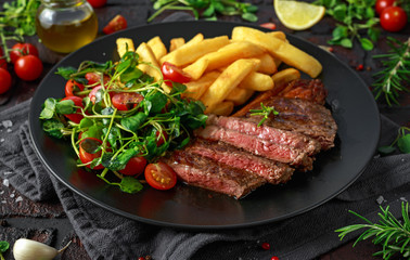 Grilled sirloin steak with potato fries and vegetables, tomato salad in a black plate. rustic table