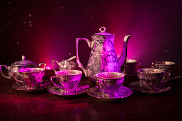 Food background tea and coffee theme. Old vintage ceramic tea or coffee pot with cups jug and sugar cup on dark background with lights and smoke. Silhouettes of ceramic tea pot and cup.