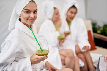 Favorite beverage. Young lady with towel on head enjoying drink and looking at camera with smile. Two women in white bathrobes and talking on blurred background