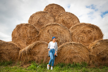 Naklejka na ściany i meble Pretty fresh darkhaired young woman walking outdoors in the sun against the background of haystacks and wearing a coat, knitting hat, jeans.Concept of autumn holidays at village and live style