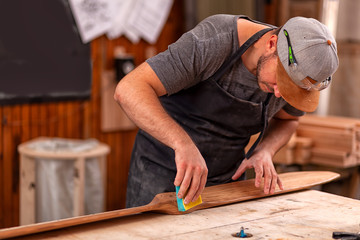 Experienced carpenter in work clothes and small business owner working in woodwork workshop,  using sandpaper for polishing children's helicopter blades at worktable in workshop