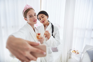 Unforgettable event. Smiling young woman in cute diadem taking photo with best friend. Ladies holding glasses with champagne
