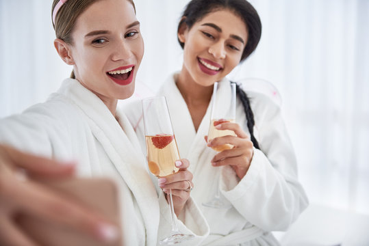 Good shot. Cheerful young woman taking photo with friend. Women holding glasses with champagne and wearing white bathrobes