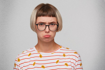 Closeup of unhappy pretty young woman wears striped t shirt and spectacles feels disappointed and curving her lip isolated over white background