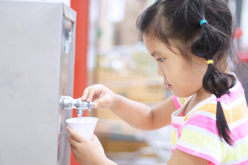 asian children cute or kid girl thirsty and press cold water from refrigerator or water dispenser for drink in white paper cone cup in kindergarten school or department store and super market