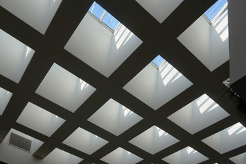 a square concrete geometric ceiling with a multitude of windows on top from where the sun shines indoors