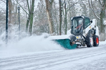 a small tractor with a rotating brush clears a wide road in the city park from the fresh fallen snow on a clear cold winter day