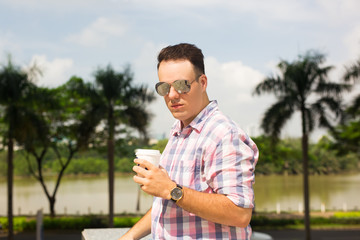Fototapeta premium Cool handsome man in sunglasses drinking coffee in summer park. Serious confident young man in casual shirt looking at camera. Modern businessman concept