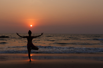 Fototapeta na wymiar Young slender girl with her back doing yoga at sunset on the beach. Pose on balance, pose tree. Hands are raised and connected together. Natural backlight.