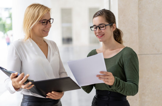 Smiling Young Manager Giving Paper Report To Friendly Boss Assistant. Two Business Employees In Glasses Reviewing Documents In Open Folder. Paperwork Concept