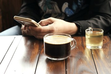 Fototapeta na wymiar Black coffee in clear glass and a hot tea served on a wooden table in front of a woman reading a text message on a mobile phone.