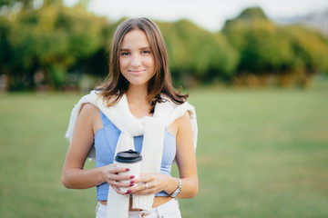 Portrait of beautiful charming woman with short dark hair, has healthy skin, dressed in casual clothes, holds takeaway coffee, enjoys recreation time, stands against blurred nature background