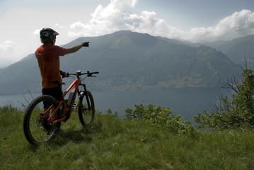 Fototapeta premium man with electric bicycle, e-bike, ebike, view of mountains, valley and lake of Como, summer, sport, adventure, cloud, sun, alps, Lombardy, Italy