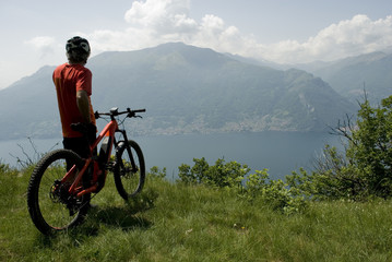 Fototapeta premium man with electric bicycle, e-bike, ebike, high mountains, observes horizon, valley, lake of Como, forest, summer, sport, adventure, cloud, sun, alps, Lombardy, technology, Italy
