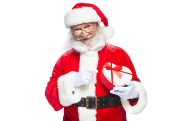 Christmas. Smiling Santa Claus in white gloves holds a red and white heart-shaped gift box with a red ribbon. Isolated on white background.
