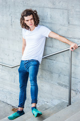 Young Hispanic American College Student thinking outside in New York, with long brown curly hair, wearing white T shirt,  blue jeans, green sneakers, standing on stairs against wall, looking away, sad