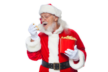 Christmas. Smiling, kind Santa Claus in white gloves with his mouth open holds a red bucket with popcorn with one hand, takes some popcorn and prepares to eat it with the second hand. The concept of