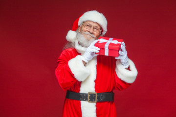 Christmas. Kind Santa Claus in white gloves holds a gift red box with a bow near an ear. He listens. Isolated on red background.