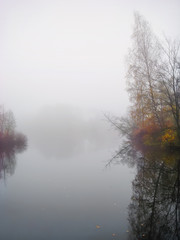 Foggy dawn, autumn forest in mystic fog in the morning, lake at dawn with clouds reflected in the calm water, thick dense fog.