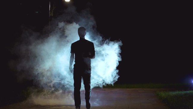 The male walking near the cloud of a smoke on the dark background, slow motion