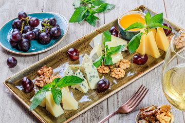 Cheese platter garnished with honey, walnuts, grapes, bread, mint and glass of wine over wooden background