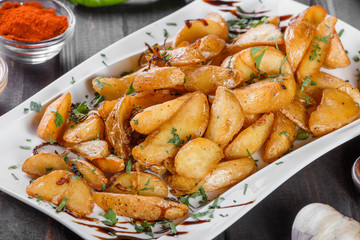 Roasted or baked potato with herbs, garlic, tomatoes and sauce on white plate on dark wooden background