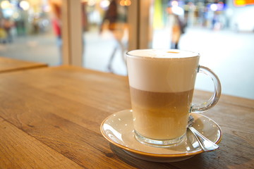 coffee latte macchiato in clear transparent glass and white plate lay on rough wooden table counter beside windows of coffee sop and blur background outside coffee shop.