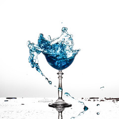A blue drink splashing out of a wine glass against a white background