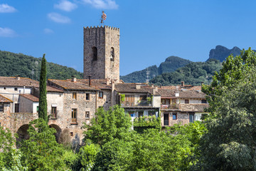 Fototapeta na wymiar Spain, Catalonia, Santa Pau: Panoramic view on the famous skyline of old ancient fortified Spanish town with tower, houses, green trees and blue sky in the background - concept travel history.