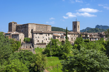 Fototapeta na wymiar Spain, Catalonia, Santa Pau: Panoramic view on the famous skyline of old ancient fortified Spanish town with tower, houses, green trees and blue sky in the background - concept travel medieval