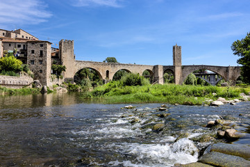 Fototapeta premium Spain, Catalonia, Besalu: Panorama view on the famous skyline of old ancient fortified Spanish town with medieval bridge, river El Fluvia, tower, houses, green trees and blue sky in the background.