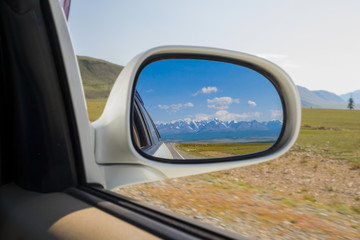Naklejka premium Reflection in the mirror of a riding white car of large altai mountains with snow on top by a sunny bright day with a blue clear sky green field and clouds