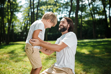 Dark-haired dad and his son dressed in white t-shirts fool around and having fun on the green lawn in the park on a sunny day