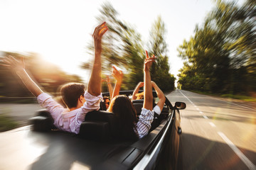 ?ompany of young people riding in a cabriolet on the road and holding their hands up on a warm...