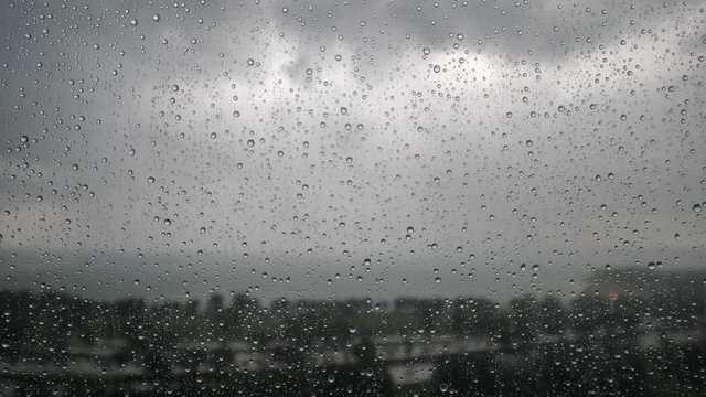 Video of rain drops collecting on a transparent glass window pane on a gloomy, cloudy and overcast, rainy weather day with focus on the droplets and panoramic view of Lake Michigan in background.
