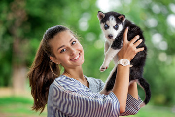 Naklejka na ściany i meble A beautiful smiling woman with a ponytail and wearing a striped shirt is cuddling with a sweet husky puppy while standing on the lawn. Love and care for pets.