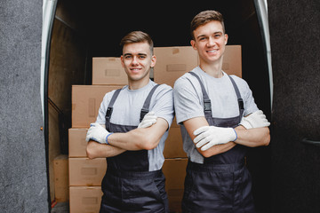 Two young handsome smiling workers wearing uniforms are standing in front of the van full of boxes. House move, mover service.